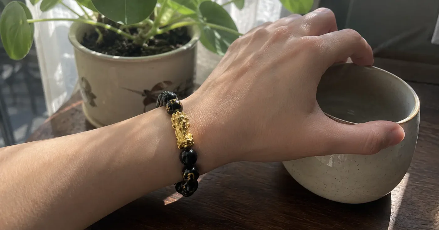 Left wrist wearing a black obsidian Feng Shui bracelet with a golden Pi Xiu charm, with the hand naturally reaching for a ceramic teacup on a wooden desk.