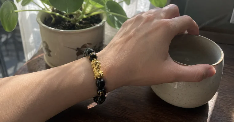Left wrist wearing a black obsidian Feng Shui bracelet with a golden Pi Xiu charm, with the hand naturally reaching for a ceramic teacup on a wooden desk.